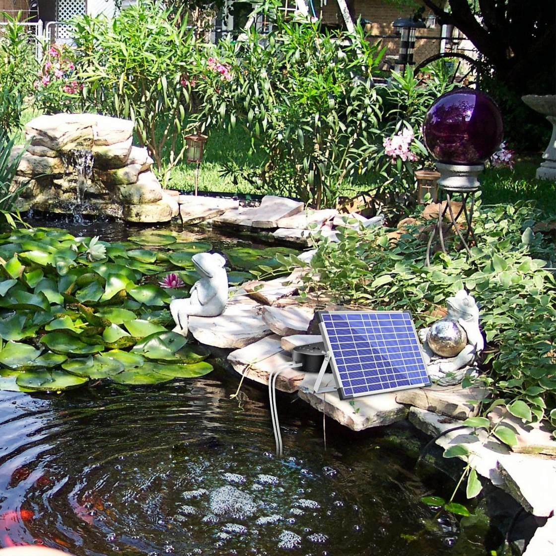a beautiful garden pond with a variety of decorative elements. There is a small waterfall cascading into the pond, surrounded by rocks and greenery. A solar panel is installed on a rock near the pond, likely powering an aerator or fountain. The pond is adorned with lily pads and a frog statue sitting on a rock. The background features lush foliage and a purple glass ball on a stand, adding to the tranquil ambiance of the garden.